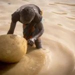Seorang anak perempuan yang bekerja di lokasi tambang emas Massakama mendulang emas dengan cangkang buah berenuk (calabash). Di wilayah Kayes di Mali, banyak anak yang kehilangan hak-hak dasar mereka atas kesehatan dan pendidikan serta bekerja di lokasi tambang emas. Kayes, 20 Maret 2019. (UNICEF/UN0293787/Keïta)